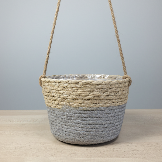 Panier tressé bicolore suspendu (moitié supérieure beige naturel et moitié inférieure gris clair), avec une doublure en plastique transparent visible, posé sur une table en bois clair.