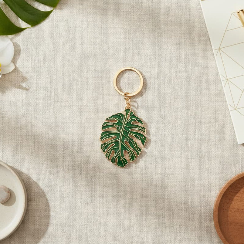 Composition à plat (flat lay) montrant le porte-clés monstera au centre d'un bureau, entouré d'une véritable feuille de monstera, d'un carnet blanc avec un stylo doré et d'éléments décoratifs naturels.