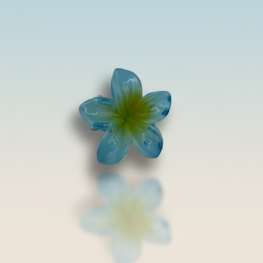 Pince à cheveux translucide en forme de fleur de frangipanier, de couleur bleue avec un centre jaune, reflétée sur une surface brillante.