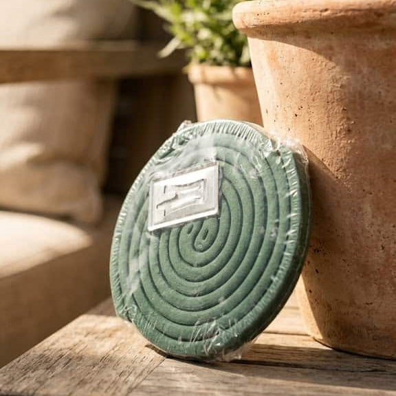Un pack de serpentins anti-moustiques verts posé sur une table en bois rustique, appuyé contre un pot de fleurs en terre cuite dans un décor de terrasse ensoleillée et chaleureuse.