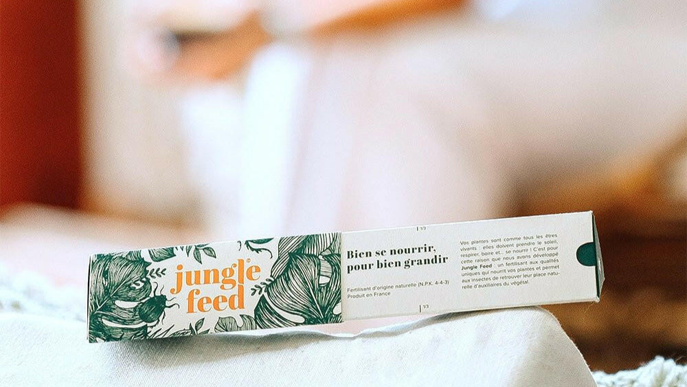 Boîte d'engrais "Jungle Feed" fermée, avec le motif de feuilles de monstera et le texte "Bien se nourrir, pour bien grandir", posée sur une nappe blanche avec des franges vertes, un arrière-plan flou.