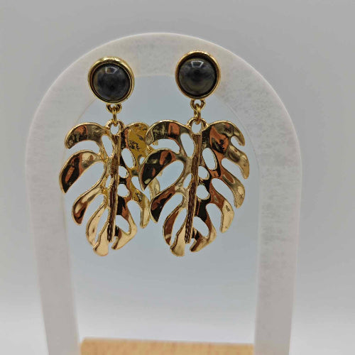 Paire de boucles d'oreilles pendantes dorées en forme de feuilles de Monstera évidées, surmontées d'un cabochon rond en pierre noire marbrée, présentées sur un support blanc.