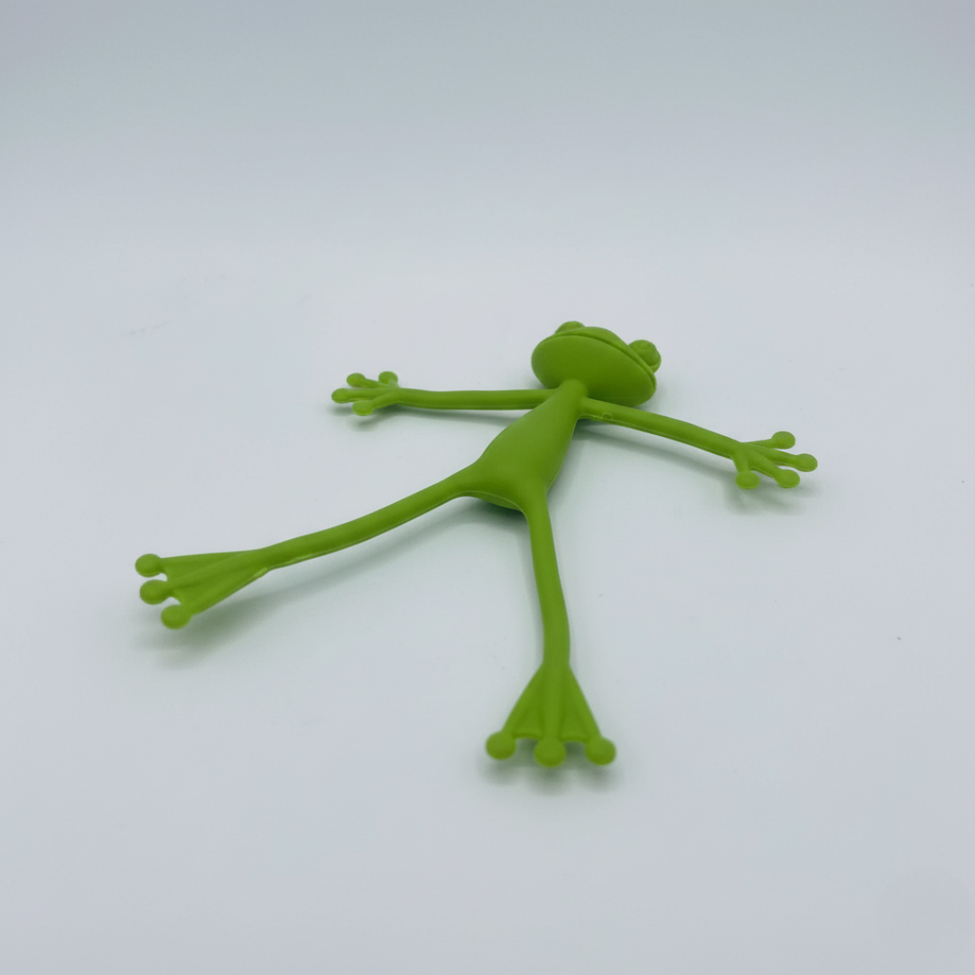Figurine souple en plastique en forme de grenouille verte avec les quatre membres et la tête écartés, posée sur un fond blanc.