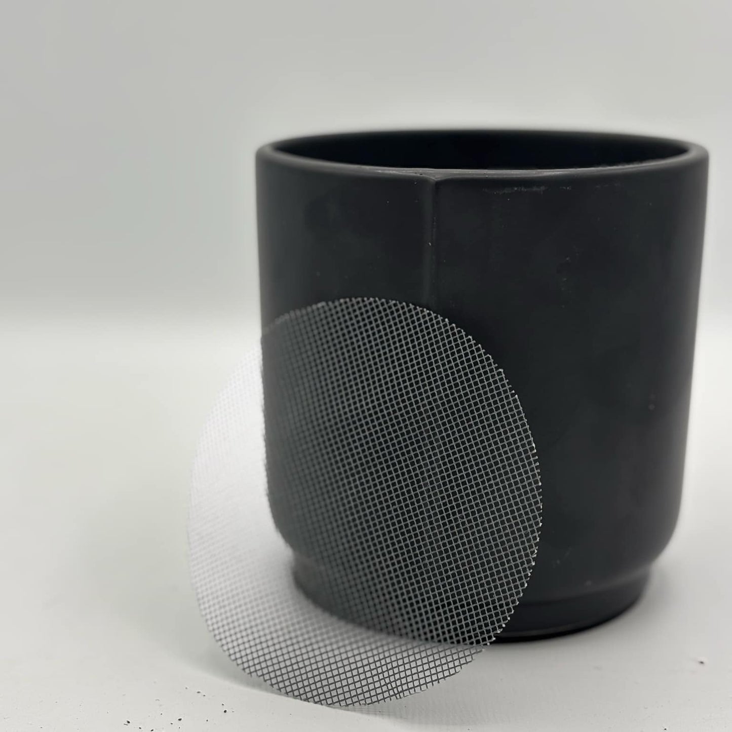 Gros plan d'une grille de drainage circulaire en plastique blanc posée contre un cache-pot noir mat sur un fond blanc neutre.