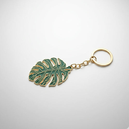 Porte-clés doré en forme de feuille de monstera avec émail vert, présenté sur un fond dégradé gris et blanc avec un éclairage de studio professionnel et des ombres douces.