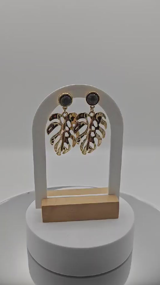 Paire de boucles d'oreilles pendantes dorées en forme de feuilles de Monstera évidées, surmontées d'un cabochon rond en pierre noire marbrée, présentées sur un support blanc.