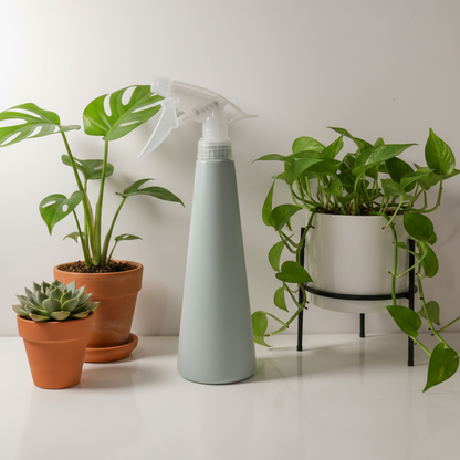 Vaporisateur pour plantes de 380 ml, bleu pâle, posé sur une surface blanche, entouré de différentes plantes d'intérieur vertes en pots (Monstera, Pothos, succulente). L'image suggère l'utilisation du produit dans un environnement végétal stylisé.